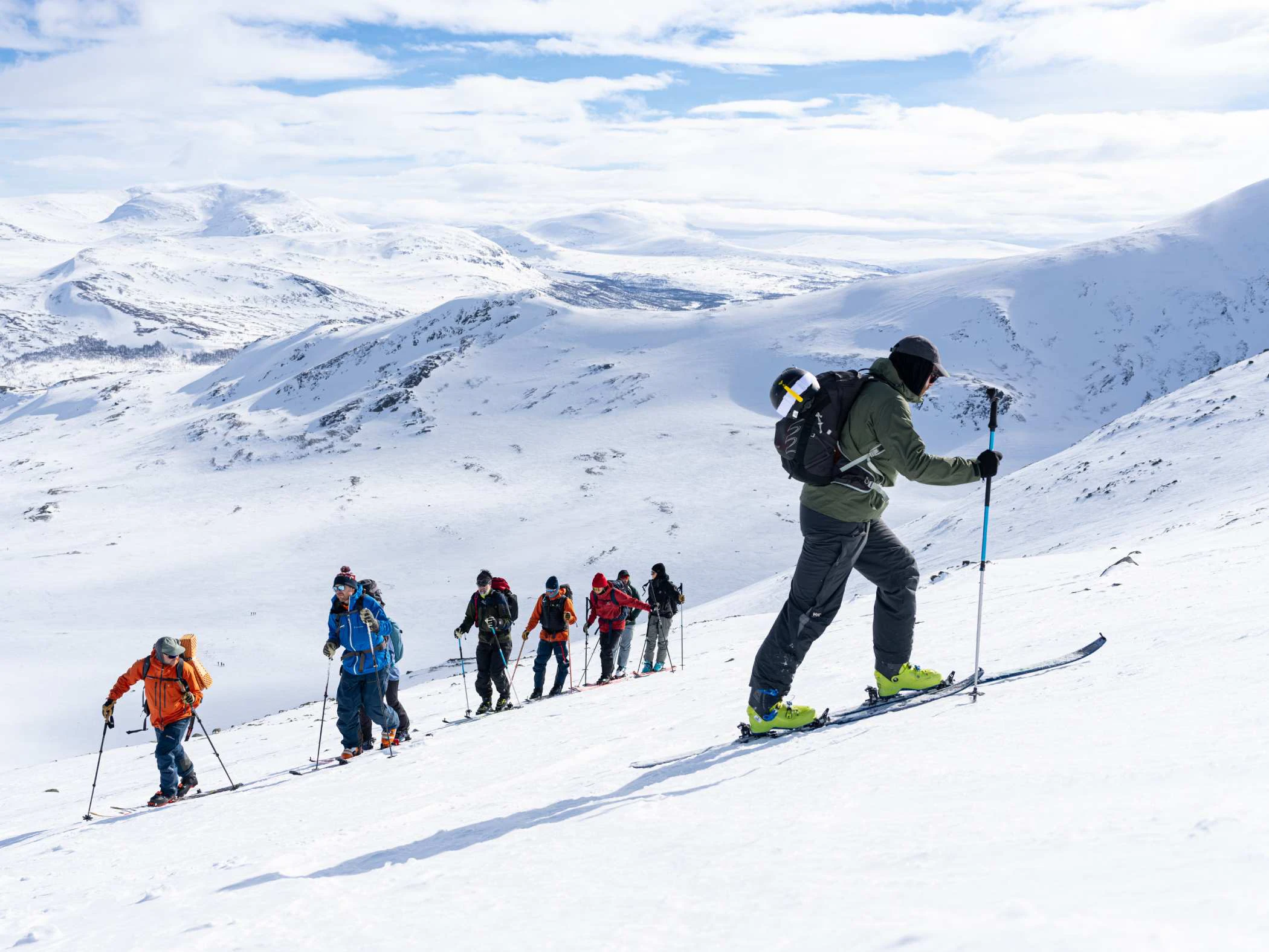 Grupp med toppturåkare på väg uppför en snötäckt fjällsluttning i vinterlandskap.
