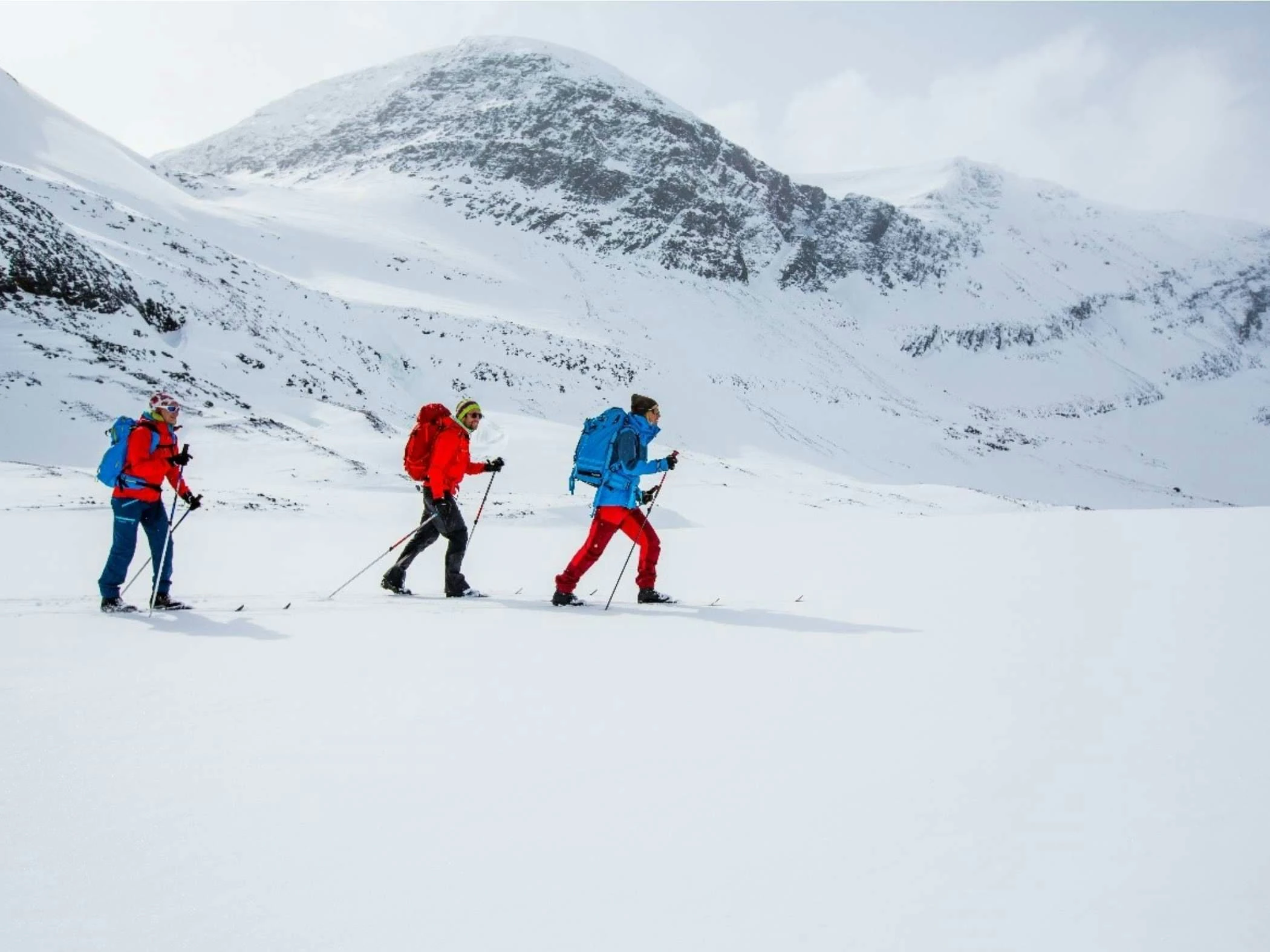 Snöklädda fjäll och tre personer på topptur med ryggsäckar och stavar i ett öppet vinterlandskap.