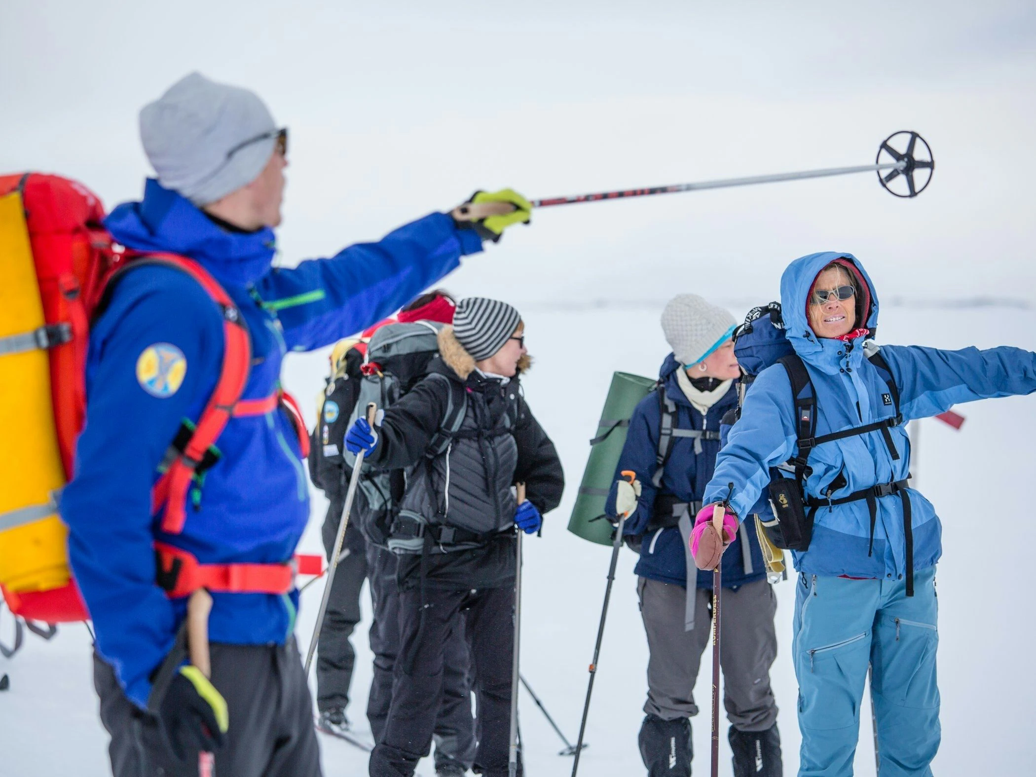 En guide visar vägen för en grupp vintervandrare med ryggsäckar och stavar i ett snötäckt fjällandskap.
