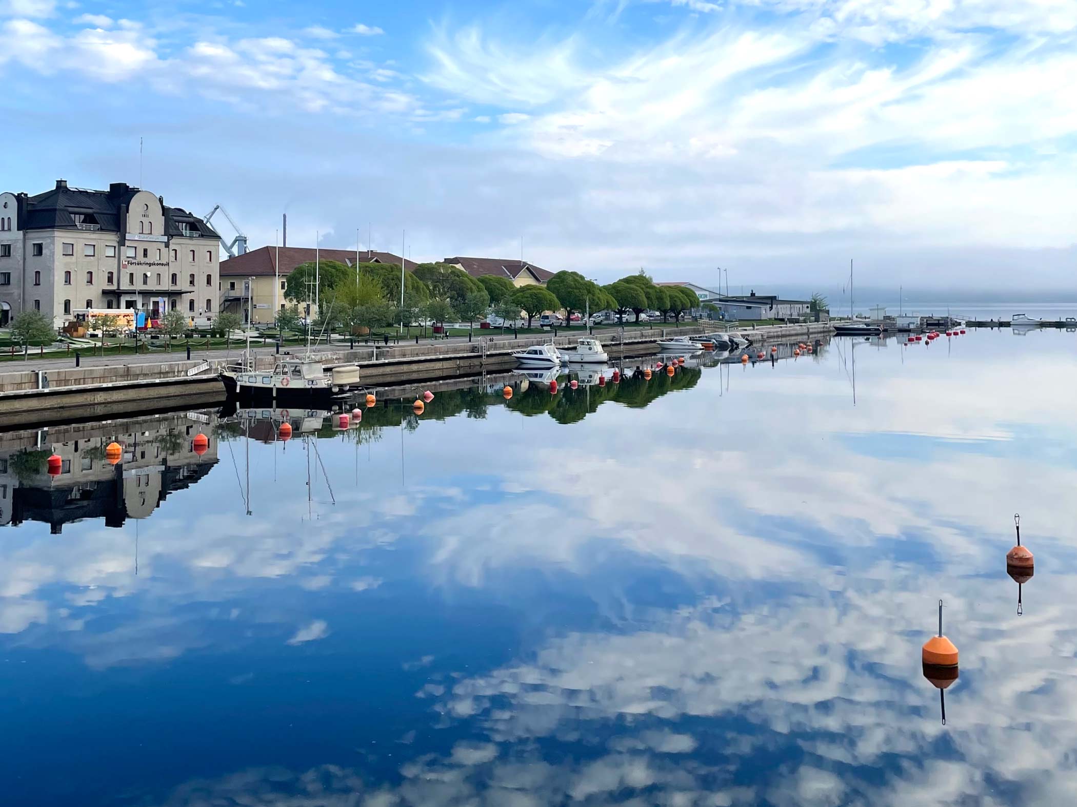 Lugn hamn med båtar och speglingar av himmel och byggnader i vattnet.