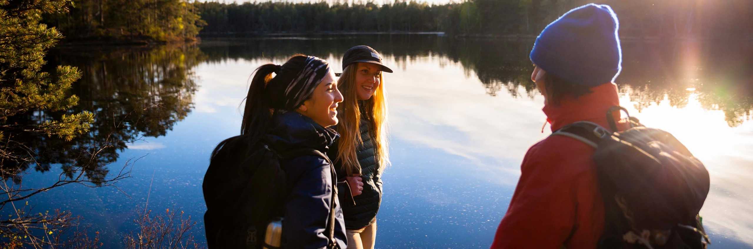 Tre personer med ryggsäckar står vid en stilla sjö i skogen och pratar i varmt kvällsljus.